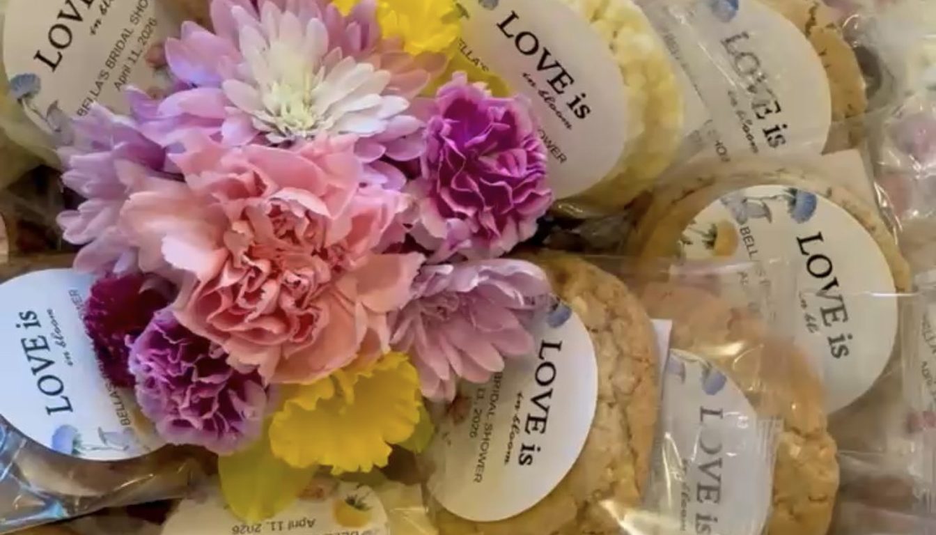 Individually wrapped cookies with custom bridal shower labels reading LOVE is in bloom, arranged around fresh flowers, from Little Miss Moffitt