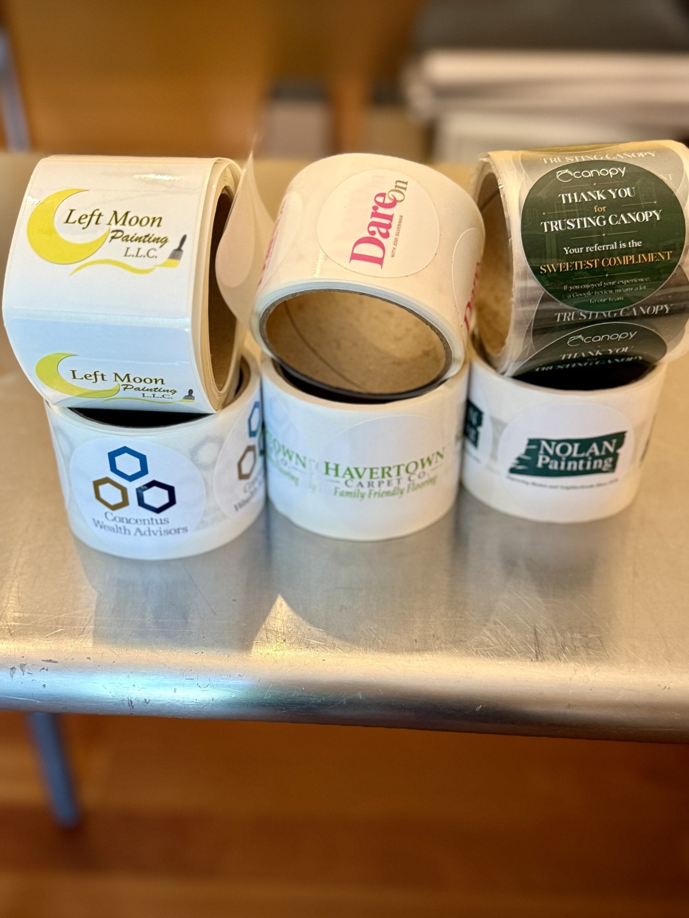 Six rolls of custom printed cookie labels, each carrying a different client company logo